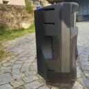 Černý reproduktor v plastovém provedení s texturovaným povrchem. Na boku jsou vidět úchyty a ventilační otvory. Nástroj stojí na kamenné dlažbě. Na f…