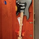Šedá elektrická kytara s černým pickguardem v oranžovém plišovém kufru. Na hlavě hředu je bílý nápis Fender a pod ním menší nápis Elite. Na těle kyta…