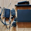 Sada černého bezdrátového systému značky SHURE. Obsahuje přijímač s nápisem SHURE, vysílač s nápisem SHURE a napájecí adaptér s nápisem SHURE PS4E. S…