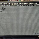 Kytarový zesilovač s černým krytím a šedou textilní mřížkou. Na horním ovládacím panelu jsou knofíky a nápisy NORMAL, VIBRATO, Twin Reverb-Amp. Na př…