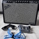 Kytarový zesilovač s černým povrchem a šedou přední síťovkou. Na horním ovládacím panelu jsou nápisy NORMAL, VIBRATO a Deluxe Reverb-Amp FENDER MUSIC…