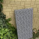 Obdélníkový akustický panel potažený látkou s černobílým geometrickým vzorem. Panel je opřen o žlutou kamennou zeď vedle zelené vegetace. Na povrchu…