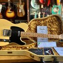 Světlý elektrický kytara typu Telecaster s černým pickguardem a javorovým hnedlem. Na hlavě je viditelné logo Fender. Kytara leží v otevřeném hnědém…