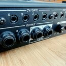 Detail zadní strany černého elektronického zařízení s řadou konektorů. Horní řada obsahuje vstupy označené jako CV/GATE (čísla 4, 3, 2, 1) a MIDI (OU…
