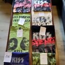 Sestet triček s motivem kapely KISS rozložených na dřevěném stole. Trička mají různé barvy (černá, žlutá, světle zelená, bílá, modrá) a potisky s čle…