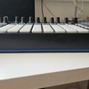 Černý MIDI kontrolér s bílými padovými tlačítky a řadou černých otočných knoflíků. Zařízení má modrý spodní rám a na levé straně je viditelný vstup p…
