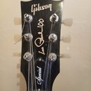 Černá hlava kytary s bílými nápisy Gibson, Les Paul 100 a Special. Jsou na ní vidět šest ladicích kolíčků s kovovými mechanismy a nylonovými knoflíky…