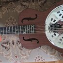 Hnědá akustická kytara typu resonator s kovovým, chromovaným krytem na těle. Na červeném zavěšeném štítku je nápis Harley Benton. Nástroj vykazuje v…