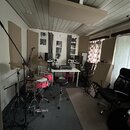 Fotografie zachycuje domácí studio s akustickými panely na stěnách a stropě. V místnosti se nachází bubnová souprava s růžovými bubny, černé studio m…
