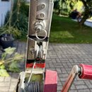 Fotografie detailu kovového mechanismu s navíjecím lanem a červenou klikou. Součásti jsou vyrobeny z kovu s viditelnými známkami opotřebení, škrábanc…