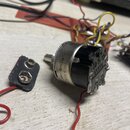 Fotografie zobrazuje elektronickou součástku, pravděpodobně potenciometr, s kovovým tělem a černým plastovým základem. Na boku je viditelný nápis M25…