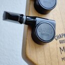 Detailní záběr na dvě černé ladicí mechanismy kytary s nápisem CHAPMAN GUITARS na knírcích. Nástroj má světle hnědý dřevěný krk, na kterém je částečn…