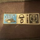 Detail štítku na hnědém textilním povrchu. Štítek obsahuje tři sekce: modrou s nápisem PROTECTION RACKET a grafickým symbolem, středovou s ikonou pos…