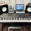 Digitální klavír šedé barvy s nápisem ROLAND DIGITAL PIANO RD-1000. Na klavíru a vedle něj jsou umístěny dva černé studiové reproduktory, stříbrný mi…