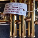 Detail zlatavého mosazného nástroje s několika ventily. Na nástroji je připevněna bílá papírová štítková visačka s nápisy: „№ ATR 313 P“, „AMATI - DE…