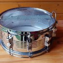 Kovový snare drum s chromovaným povrchem, umístěný na dřevěném stole. Na horní bláně je viditelné logo EVANS a přes ni je napnuté kovové sítko. Nástr…