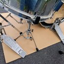 Dvě chromované pedály značky TAMA s nápisem THE LEGEND IN INNOVATION. Pedály jsou umístěny na hnědé podložce pod chromovaným bubnem. Povrch pedálů vy…