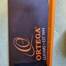 Černý obálkový nebo ochranný obal s oranžovým okrajem. Na povrchu je oranžový nápis ORTEGA® GUITARS • EST 1994 a stylizované logo v podobě velkého pí…