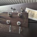 Detail hlavy akustické kytary z tmavého dřeva s kovovými ladičkami. Na dřevě je vyryté logo Sigma Guitars a nápis EST 1970. K hlavě je připevněna pap…