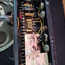 Fotografie zachycuje vnitřek elektronického zařízení s viditelnou zapojovací deskou, kondenzátory a jednou elektronovou válvou. Na jednom z komponent…