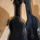 Otevřený černý tvrdý kufr na kytaru s vnitřním čaloupřením z černého plyšového materiálu. V horní části je integrovaný úložný prostor pro příslušenst…