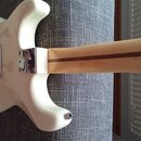 Zadní strana bílé elektrické kytary s jasně viditelným vzorem květin v laku. Na spojení krku a těla je kovový kryt s nápisem Fender. Krk kytary je vy…