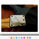 Detail krku kytary s černým lakem a kovovým krytem v místě spoje s hřidlem. Na kovovém krytu je vyryto logo Fender®. Povrch kytary vykazuje drobné od…