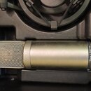 Kondenzátorový mikrofon v matné stříbrno-šedé barvě s kovovou síťkovou hlavicí. Na těle je vpravo nápis Made in Australia. Mikrofon leží v černé pols…