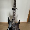 Černá elektrická kytara s tmavým dřevěným hředníkem a dvěma humbuckery. Na hlavě je zlatý nápis Ibanez. Kytara má na hředníku bílé ozdobné vložky a j…
