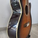 Akustická kytara s tmavě hnědým přechodem do žlutého odstínu (sunburst) a bílým lemováním hran. Na boku je umístěn černý ovládací panel s nápisem VOL…