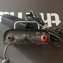 Černý přenosný zesilovač s kovovou rukojetí. Na zadní straně jsou nápisy PHONES / LINE, DC 18 V, SPEAKER OUTPUT a IMPEDANCE s hodnotami 4, 8, 16. Pří…