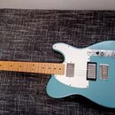Modrá elektrická kytara s bílým pickguardem a světlou hnedou hmatníkovou deskou. Na hlavě je viditelné logo Fender. Kytara má dva snímače a kovový mo…