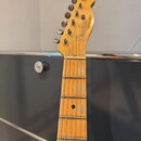 Hlava a hриф elektrické kytary z jasného dřeva. Na hlavě je černé logo Fender a nápis TELECASTER v rámečku. Hриф má viditelné opotřebení laku a značk…