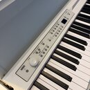 Bílá digitální klaviatura s černými a bílými klávesami. Na ovládacím panelu je viditelné logo KORG, regulační knoflík VOLUME a řada tlačítek s nápisy…