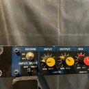 Detailní pohled na přední panel rackového efektového zařízení s modrým pozadím. Jsou zde viditelné nápisy REVERB, INPUT, OUTPUT a MIX, přepínač ON/OF…
