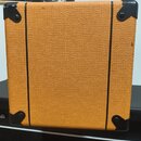 Záběr na zadní stranu kytarového komba potaženého oranžovým texturovaným vinylem. Rohy jsou chráněny černými plastovými nárožníky a povrch je prořízn…