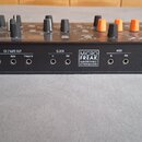 Černý syntetizátor s oranžovými a černými knoflíky. Na zadní straně jsou nápisy ARTURIA, _The sound explorers a MICROFREAK ALGORITHMIC SYNTHESIZER. J…