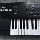 Černý klavír s bílými a černými klávesami. Na levé straně je bílý nápis Roland JUNO-D. Panel obsahuje ovládací prvky s popisky VOLUME, MODE a EDIT, k…