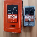 Kytarový efekt v stříbrném kovovém šasi a doprovodná oranžová krabice. Na obou je nápis SMALL STONE EH4800 PHASE SHIFTER a logo El nano s textem MADE…