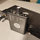 Černý bezdrátový vysílač s ovládacími prvky na horní straně. Na přední straně jsou nápisy POWER/PAR, OUTPUT, VOLUME a OFF/ON oraz kulatá nálepka s te…
