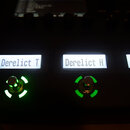 Detailní záběr na ovládací panel černého elektronického zařízení. Jsou vidět tři podsvícené displeje, z nichž první dva mají nápisy „Derelict T“ a „D…