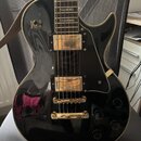 Černá elektrická kytara s gold hardwarem a krémovým lemováním. Na těle jsou dva humbuckery, čtyři ovládací knofíky a přepínač s nápisy RHYTHM a TREBL…