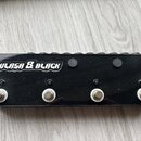 Černý obdélníkový pedalboard s nápisem ULASA & BLACK v bílém stylizovaném písmu. Na přední straně jsou čtyři kovové spínací tlačítka s bílými podložk…