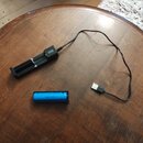 Na dřevěném stole je černý nabíjecí adaptér s USB konektorem a jedna modrá baterie. Na boku adaptéru je viditelné bílé logo, které je však nečitelné.…