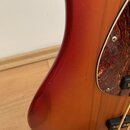 Detail těla elektrického nástroje v červeném gradientním lakování. Nástroj má hnědý vzorovaný pickguard s viditelnými šrouby. Na lakovaném povrchu js…