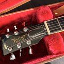 Černá hlava elektrické kytary s nápisy Gibson, MODEL Les Paul. Nástroj je umístěn v červeně vystlaném kufru, na kterém je hnědý kožený pásek s logem…