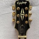 Hlava kytary značky Epiphone s černým povrchem a zlatými ladičkami. Uprostřed je umístěna bílá perleťová intarzie ve tvaru kosočtverce. Na spodní čás…