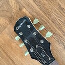 Černá hlava kytary s bílým nápisem Epiphone a označením SG. Na hlavě jsou viditelné kovové ladičky s bílými plastovými knoflíky. Povrch je černý, v h…