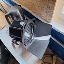Černý LED reflektor s nastavitelnými klapkami (barn doors) umístěný na bílém povrchu. Zařízení má žebrovaný chladič a v centru kulatou matici s více…