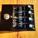 Černý kytarový efekt s nápisem OVERDRIVE a S 808. Na horní straně jsou čtyři černé otočné ovladače s popisky DI MODE, DRIVE, TONE a LEVEL. V levé čás…